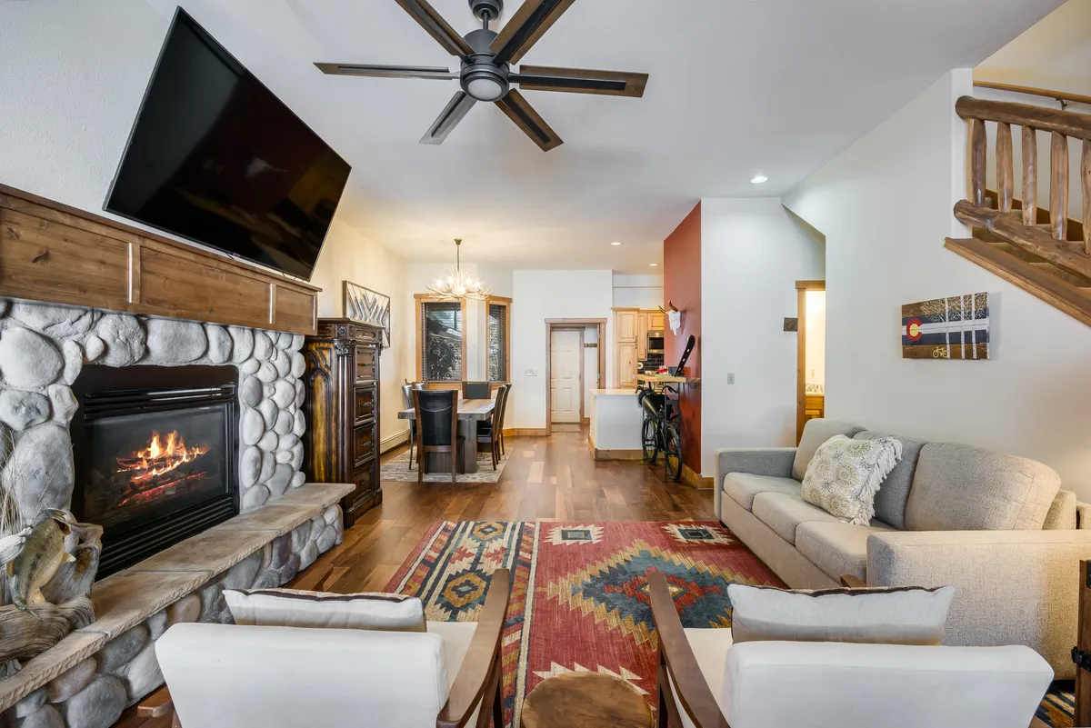 The image showcases a warm, inviting living and dining space with a glowing fireplace, plush sofa, stylish rug, and a welcoming dining table—all set beneath a ceiling fan. The room exudes comfort, perfect for gathering with family or friends, relaxing after a day on the slopes, or enjoying a cozy night in.