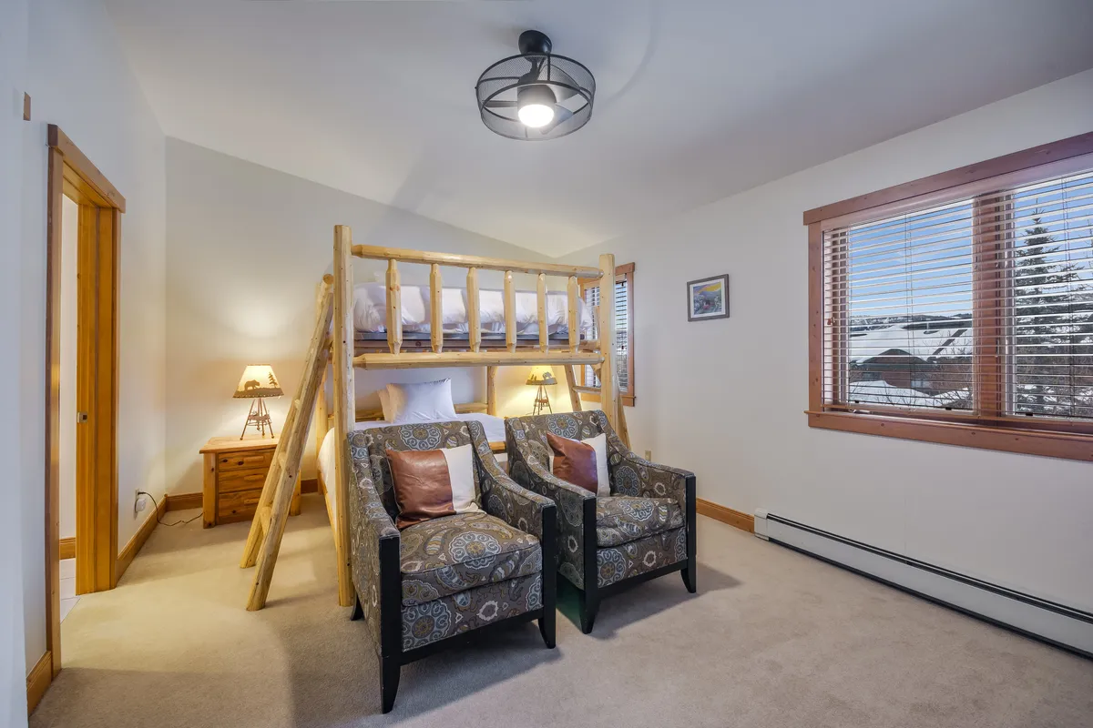 A cheerful bedroom with a queen-over-queen bunk bed, plush armchairs, and a large window offering outside views. The arrangement creates a welcoming environment for both children and adults, ideal for reading, relaxing, or family bonding before bedtime.