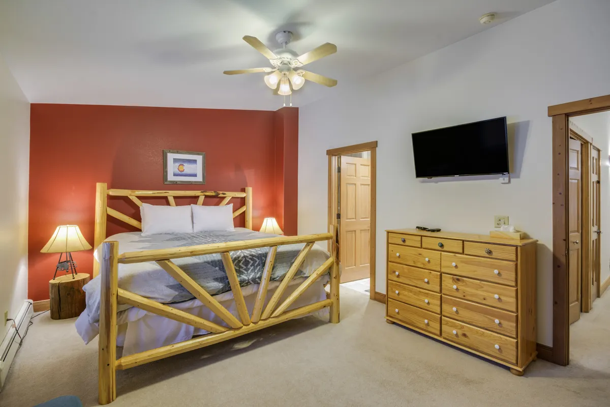 A warm, inviting bedroom featuring a wooden bed frame with plush bedding, a striking red accent wall, matching nightstands with lamps, and a wall-mounted TV. Sunlight filters softly into the room, creating a cozy ambiance ideal for relaxation or watching TV. The overall setup suggests a primary suite designed for comfort and family needs.