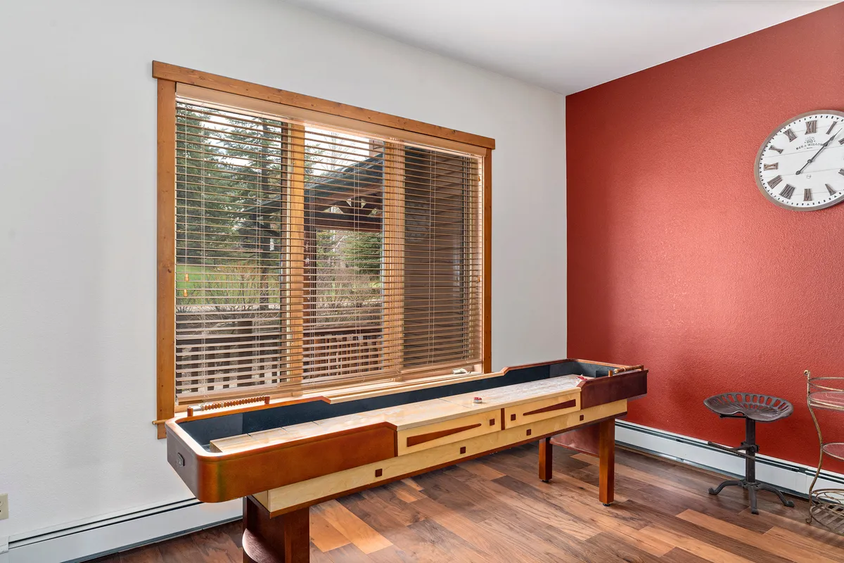 A vibrant game area with a shuffleboard table situated near a large window that lets in plenty of natural light. The red accent wall and decor create a playful, relaxed atmosphere ideal for friendly competitions and family game nights.