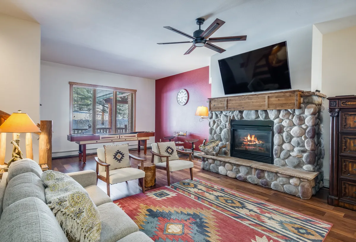 A spacious, warm living area featuring a stone fireplace, large TV, colorful rug, and wooden furnishings. The space is set up for relaxation, socializing, and games, creating a hub for family gatherings after a day of outdoor activities.