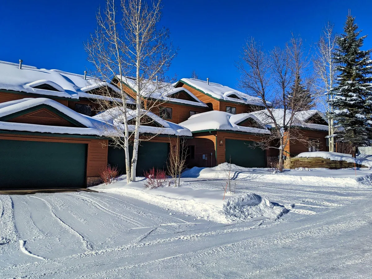 This image shows the exterior of the property with snow-covered roofs, colorful garage doors, and aspen trees framing the building. The garage doors are prominent and clearly part of the property's practical features, such as vehicle parking and gear storage.